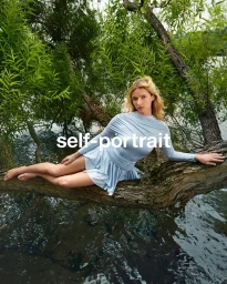 Apple Martin reclining on a large tree branch over water wearing a blue long-sleeve dress for Self-Portrait