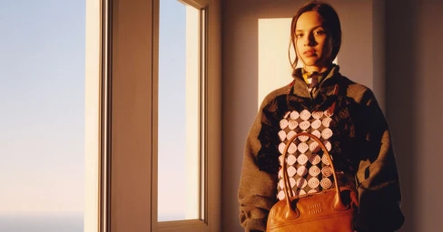 Young woman standing near a window holding a brown leather handbag, wearing a patterned top and grey sweater.