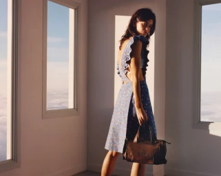 Woman in floral dress with ruffled straps holding a dark brown handbag inside a room with tall windows overlooking clouds.