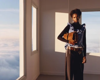 Model standing indoors by large windows overlooking clouds wearing dark trousers and a ruffled blue patterned top holding a brown leather bag