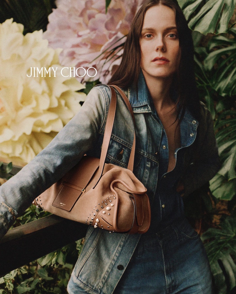 Kiki Willems wearing a denim jacket and holding a tan suede studded shoulder bag in front of oversized flowers and lush foliage.