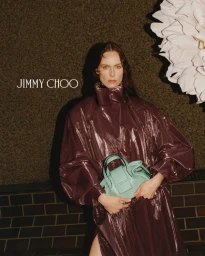Kiki Willems posing with a mint green handbag in a glossy burgundy coat next to an oversized white flower for Jimmy Choo.