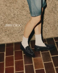 Close-up of model's legs wearing denim shorts, white socks, and black leather loafers against a tiled floor and textured stone wall