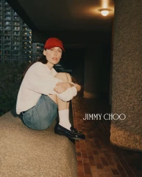 Kiki Willems wearing a red cap, white sweater, denim skirt and loafers seated outdoors for Jimmy Choo Les Fleurs project