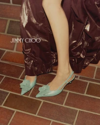 Close-up of sage green slingback heels with floral appliques worn with a deep burgundy skirt standing on a tiled brick floor.