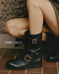 Close-up of a model wearing black leather buckled boots and a textured skirt, kneeling against a brick wall.