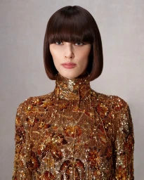 Model Mariacarla Boscono in a gold sequined high-neck top with a dark bob haircut against a neutral grey background