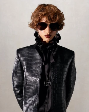 Model Mariacarla Boscono in a black textured jacket with a ruffled collar and sunglasses against a plain grey backdrop