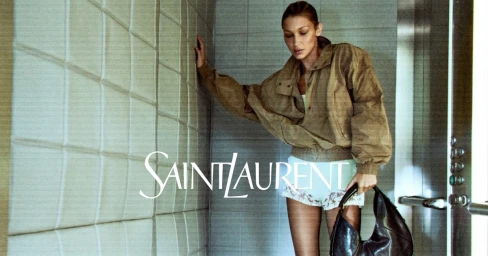 Bella Hadid leaning against a padded wall in an elevator wearing an olive jacket and white lace shorts while holding a black leather bag