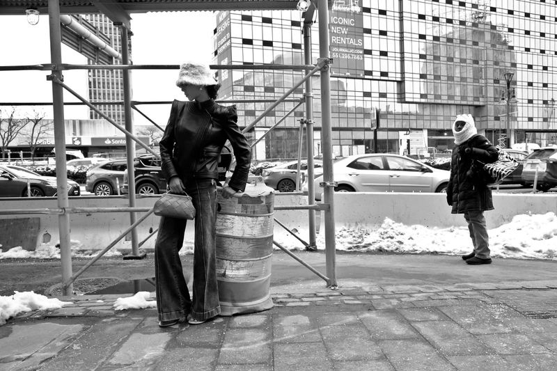 Two figures in an urban setting: one in stylish jacket leaning against a metal barrel, another fully covered near scaffolding and buildings.