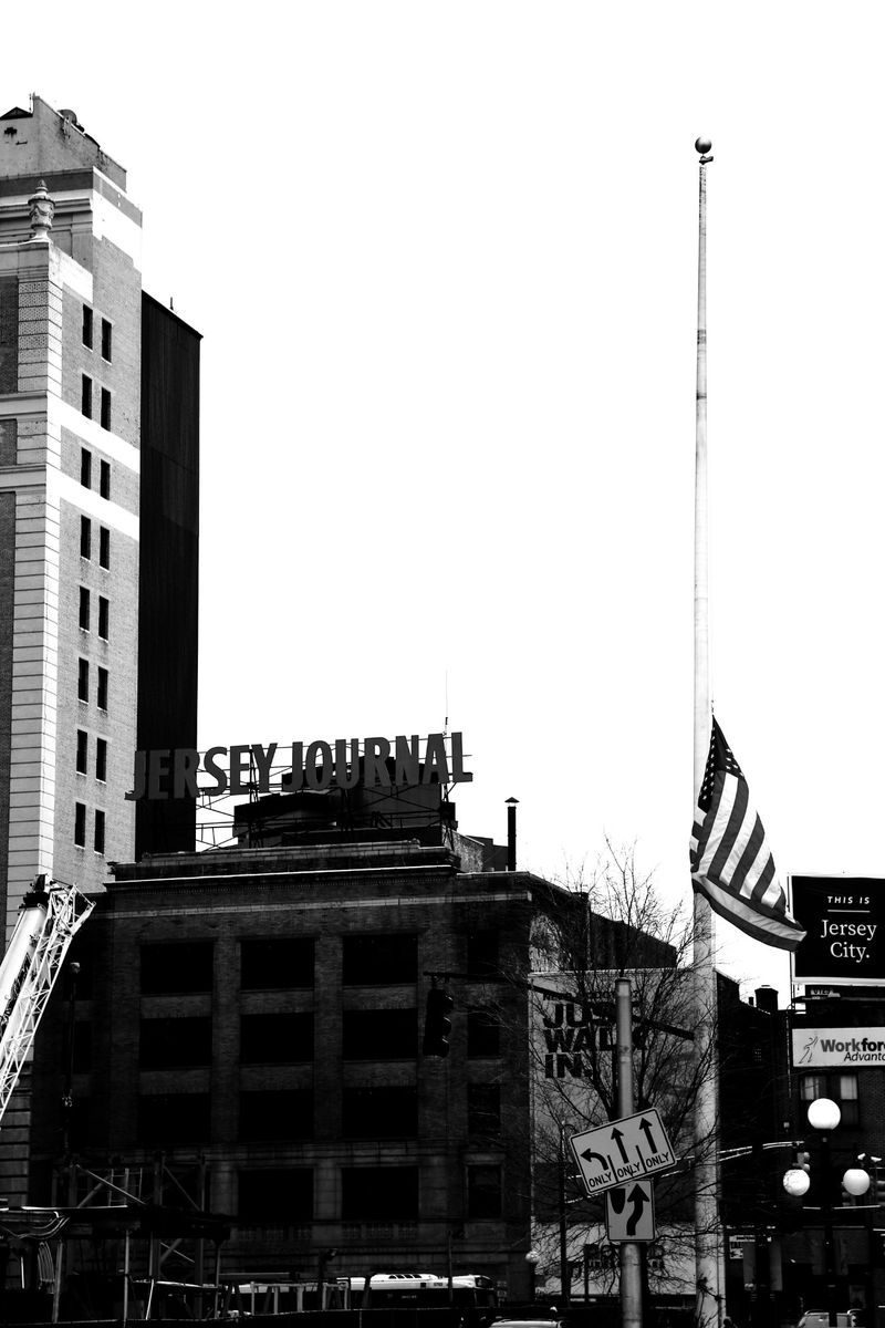 A black and white photo of the Jersey Journal building with an American flag at half-mast on a pole in an urban setting.