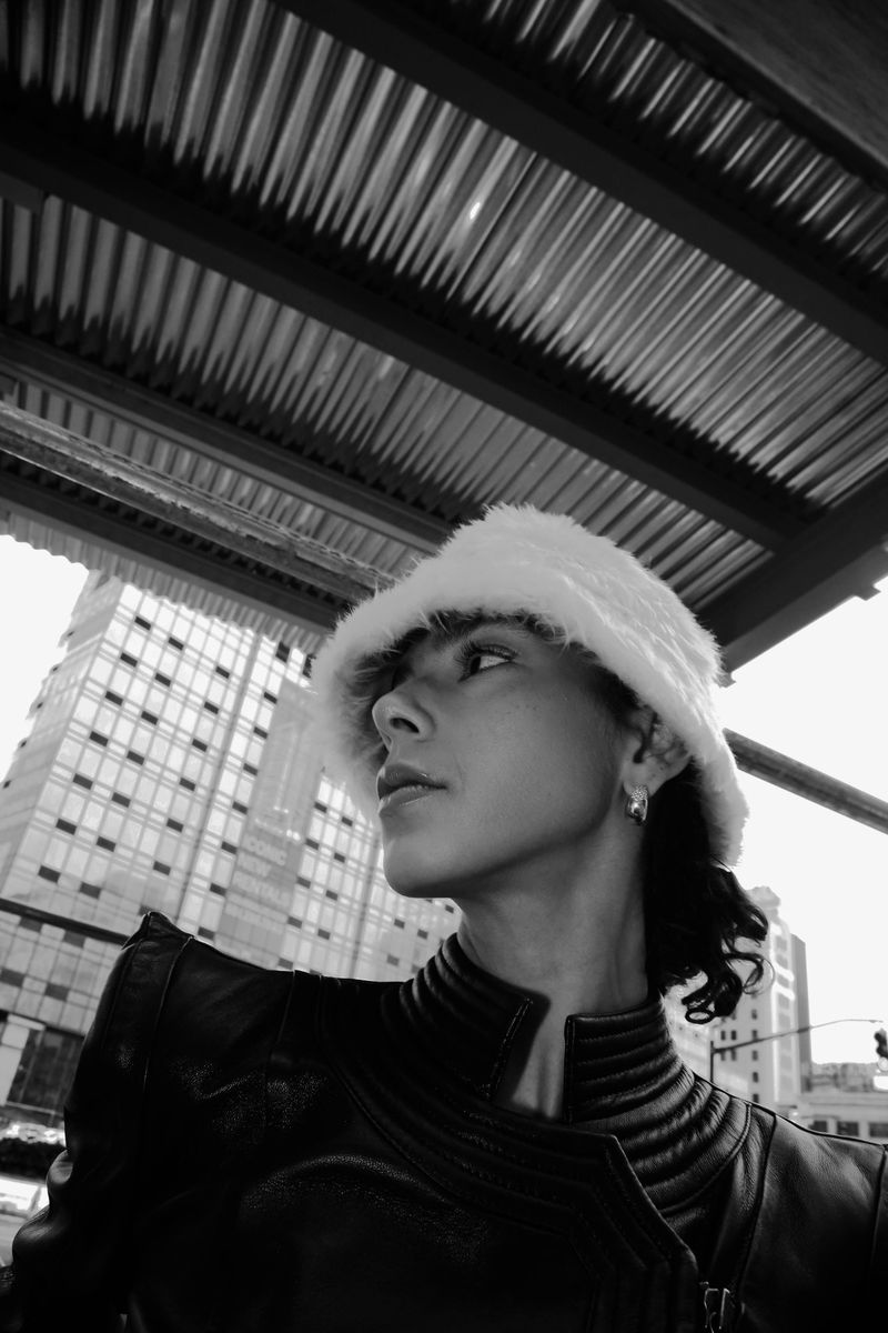A woman poses in an outdoor urban setting, wearing a fur hat and leather jacket, with a tall building visible in the background in monochrome.