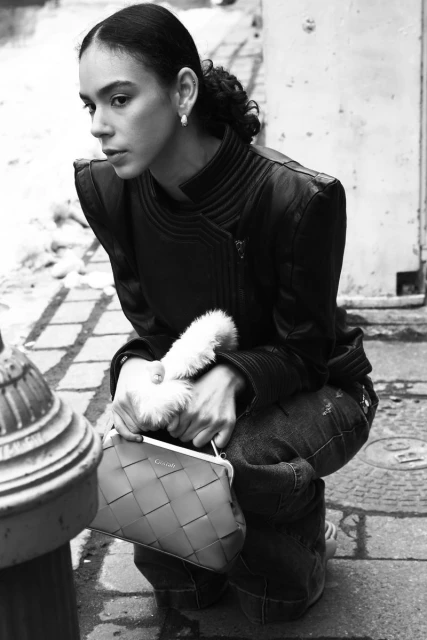 Black and white shot of a woman crouching on a city street holding a woven bag, wearing a leather jacket, and jeans.