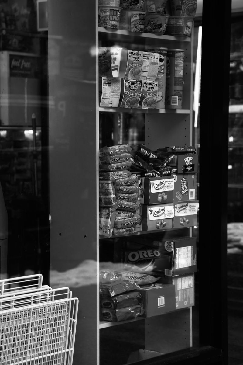 Black and white image of a shop shelf stocked with packaged food items, including snacks and breakfast foods, behind a glass window.