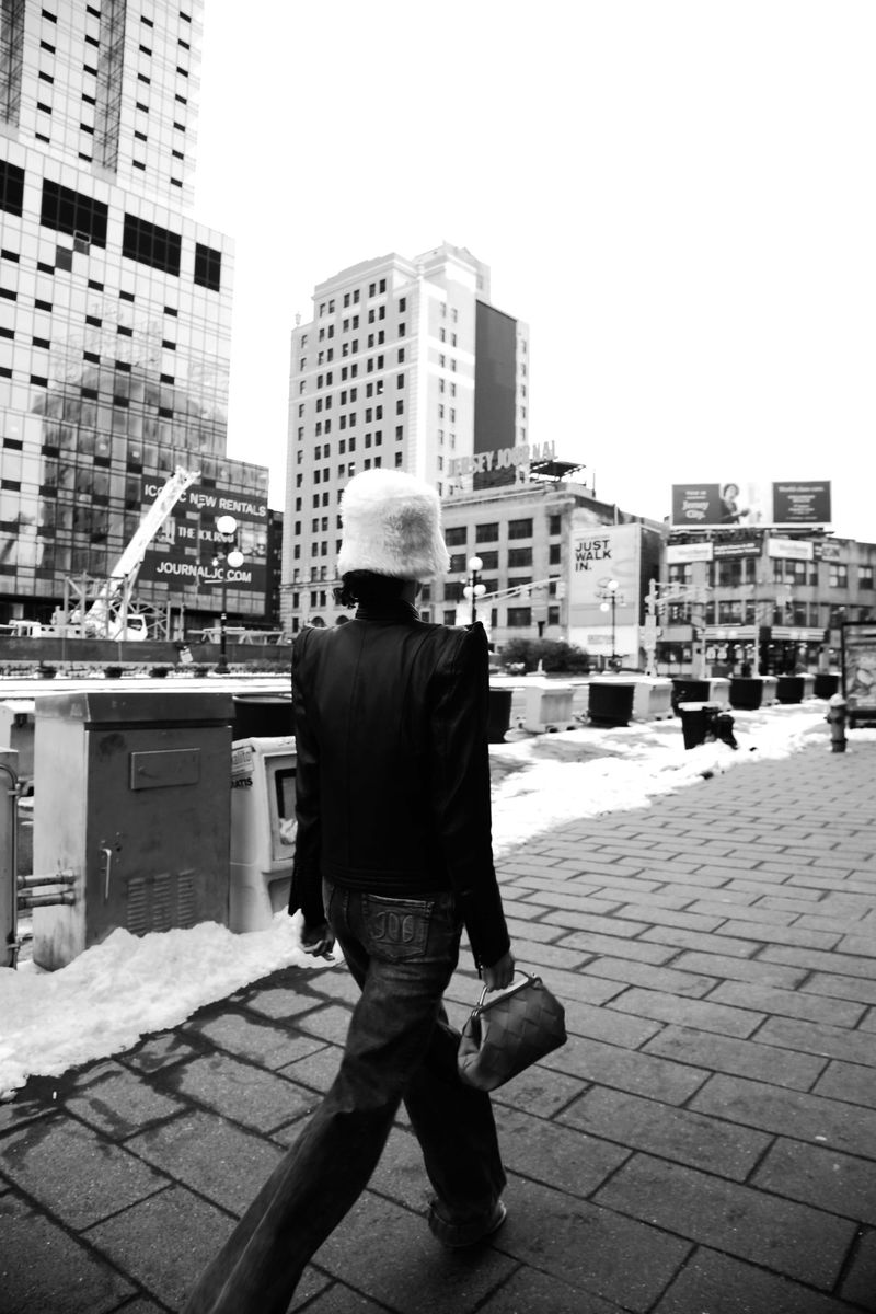 A person walks on a city sidewalk in a black and white outdoor shot, wearing a distinctive hat, jacket, and jeans.