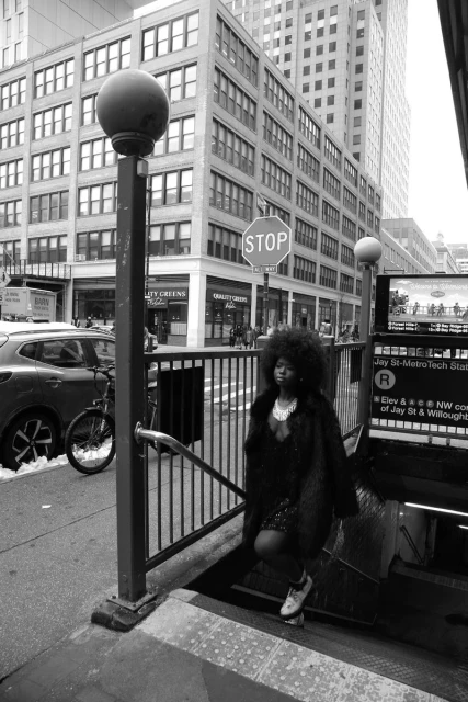 A young woman with dark curly hair in a coat standing near the entrance to the Jay St-MetroTech subway station in New York City.