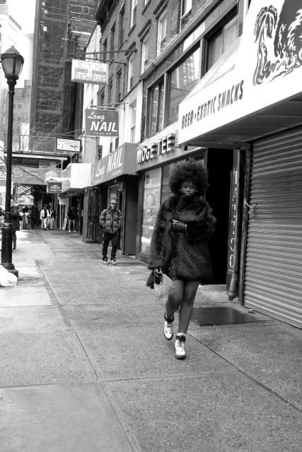A woman with an afro and fur coat walks on a city sidewalk past storefronts on a cloudy day, captured in black and white.