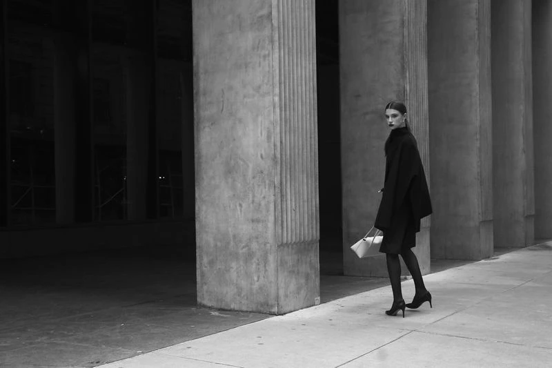Black and white full shot of a stylish woman in a dark coat carrying a handbag walking past the grand columns of a building.