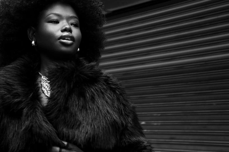 Black and white portrait of a Black woman with an afro wearing a fur coat and jewelry, posed in front of a roll-up metal door.