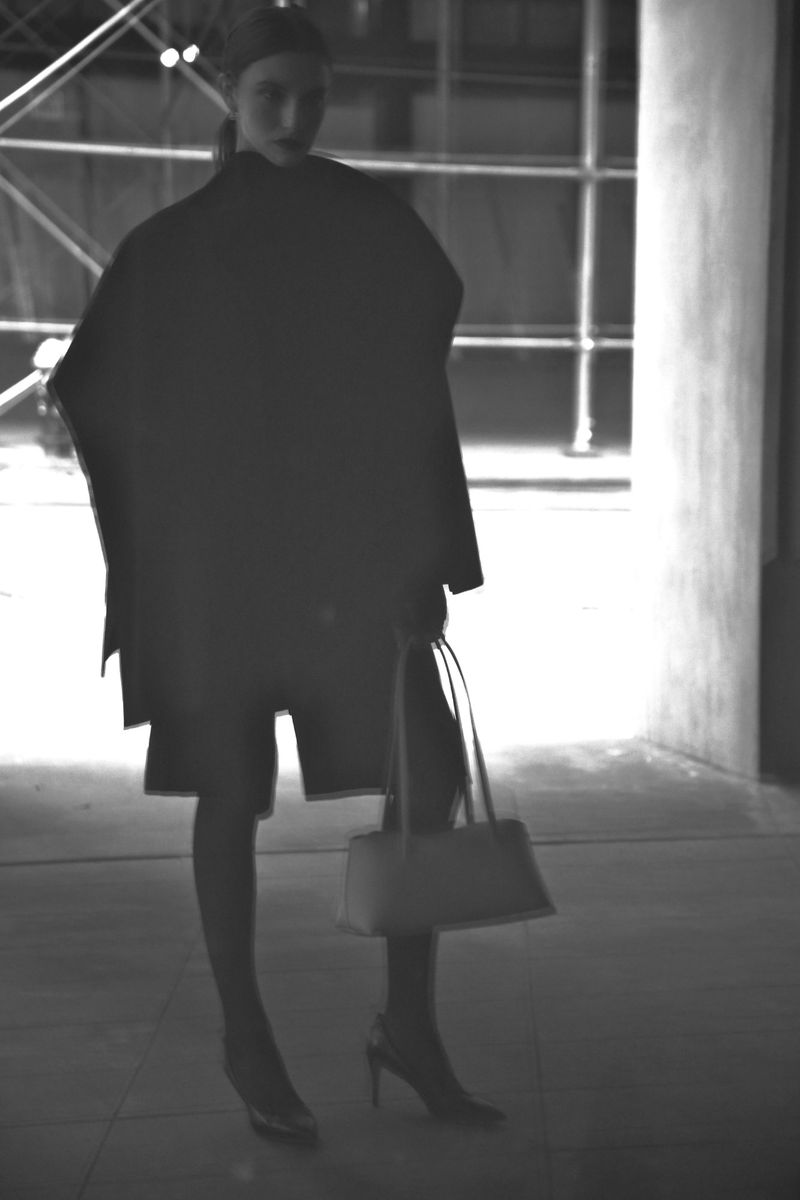 Monochrome fashion shot of a woman in an oversized jacket and heels holding a handbag near scaffolding.