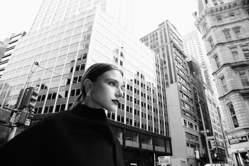 Black-and-white portrait of a woman with dark lipstick in front of modern skyscrapers in an urban environment with natural lighting.