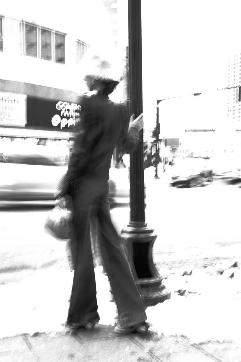 Black and white long exposure captures a blurred woman walking on a city sidewalk past a lamp post and shops on a sunny day.