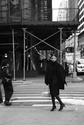 Woman in black coat holding light handbag standing on a city street crossing under scaffolding in a monochrome fashion editorial