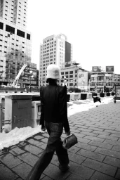 Monochrome fashion photograph of a woman walking away on a city sidewalk wearing a dark jacket, jeans, fluffy hat, and holding a small handbag