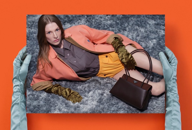 Gloved hands holding a photo of Hunter Schafer reclining on a grey rug with a leather handbag against a bright orange background.