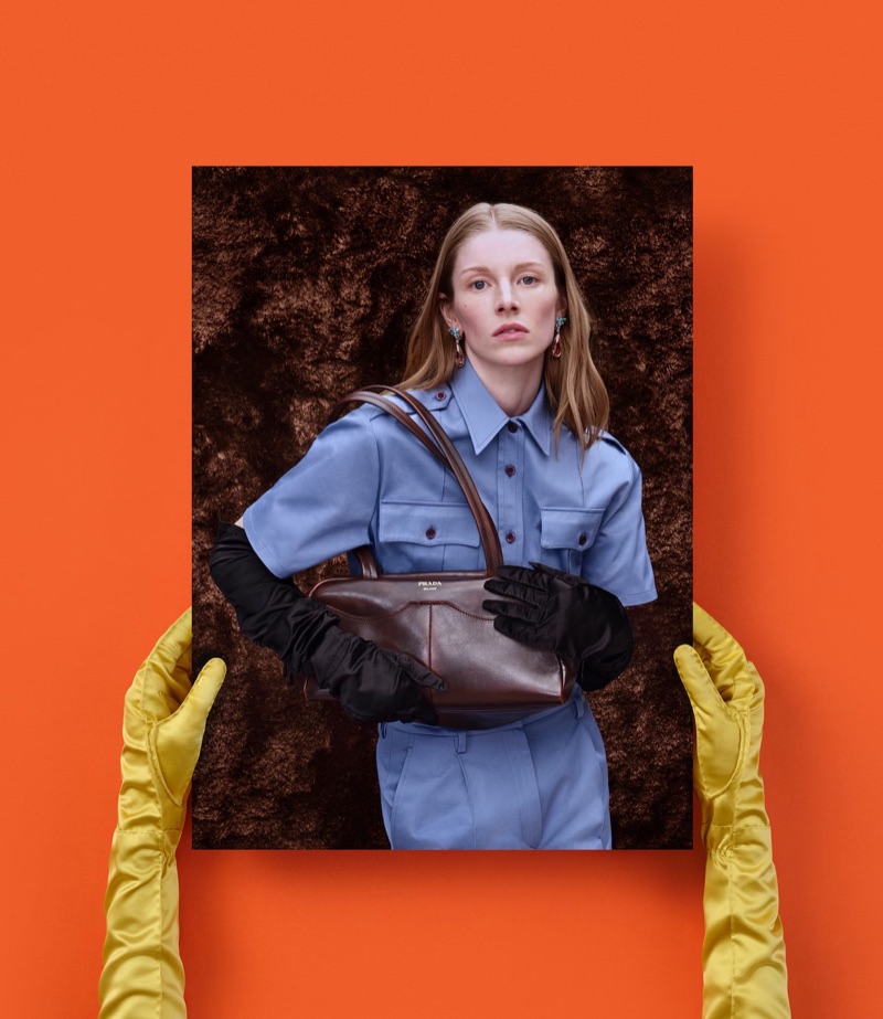 Gloved hands in yellow satin holding a photographic print of Hunter Schafer with a brown Prada handbag against an orange backdrop.