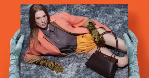 Model lying on a grey shaggy carpet wearing an orange jacket, grey shirt, and yellow shorts with dark brown handbag
