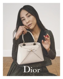 Greta Lee posing for Dior holding a light handbag against a plain background