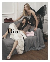Two models posing in an interior room with a grey sofa, one standing in a black lace dress and one seated wearing a white dress