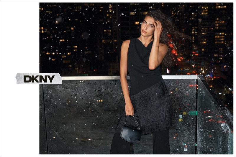 Woman wearing a black sleeveless top, fringe overlay, and trousers, holding a black handbag on a balcony overlooking a night city skyline.