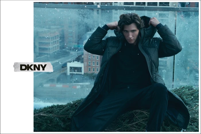 Man wearing a dark puffer coat holding the hood over his head on a snowy balcony with a city view for DKNY