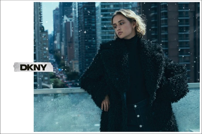 Model in a black fuzzy coat leaning against a railing with city skyscrapers and snow falling in the background