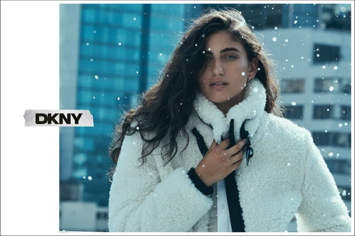 Model wearing a white fleece jacket against a city background with falling snow for the DKNY Holiday 2025 campaign.