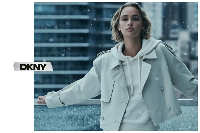 Model wearing a light jacket and hoodie outdoors against a blurred building background with falling snow for DKNY.