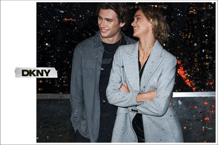 A man and a woman smiling outdoors at night amidst falling snow, wearing layered winter apparel for DKNY.