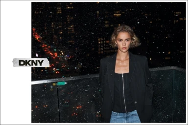 Model wearing a dark blazer and zipped top against a nighttime urban rooftop background with city lights and falling snow.