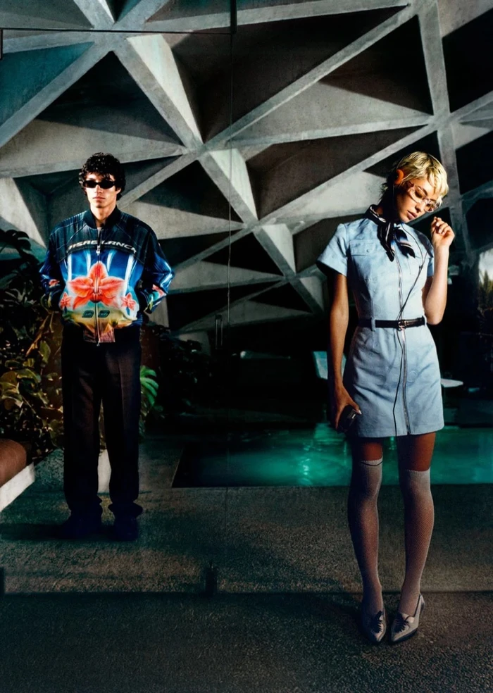 Model Iris Law in light blue uniform dress standing near indoor pool beneath geometric concrete ceiling by Corentin Leroux for Casablanca.