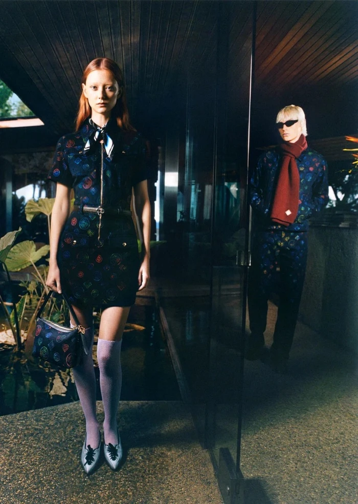 Model Iris Law standing indoors holding a printed handbag, wearing a printed dress and lilac knee-high socks, with another person in background