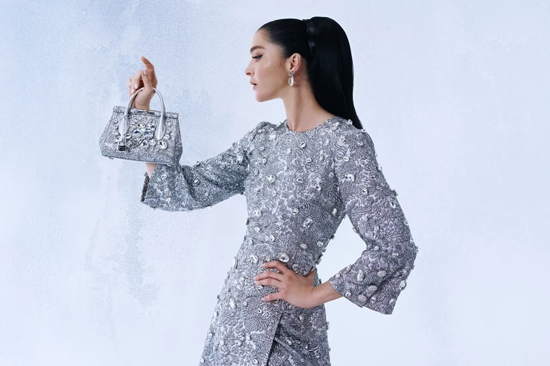 Model in embellished silver dress holding a crystal handbag against a pale textured background for Dolce & Gabbana Holiday 2025.