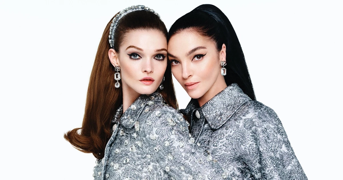 Two models wearing embellished silver jackets and statement earrings posed together against a white seamless background for Dolce & Gabbana