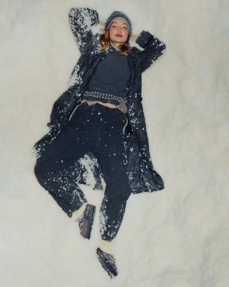 Gigi Hadid lying in snow wearing a dark grey knit coat, sweater, trousers, beanie and loafers.