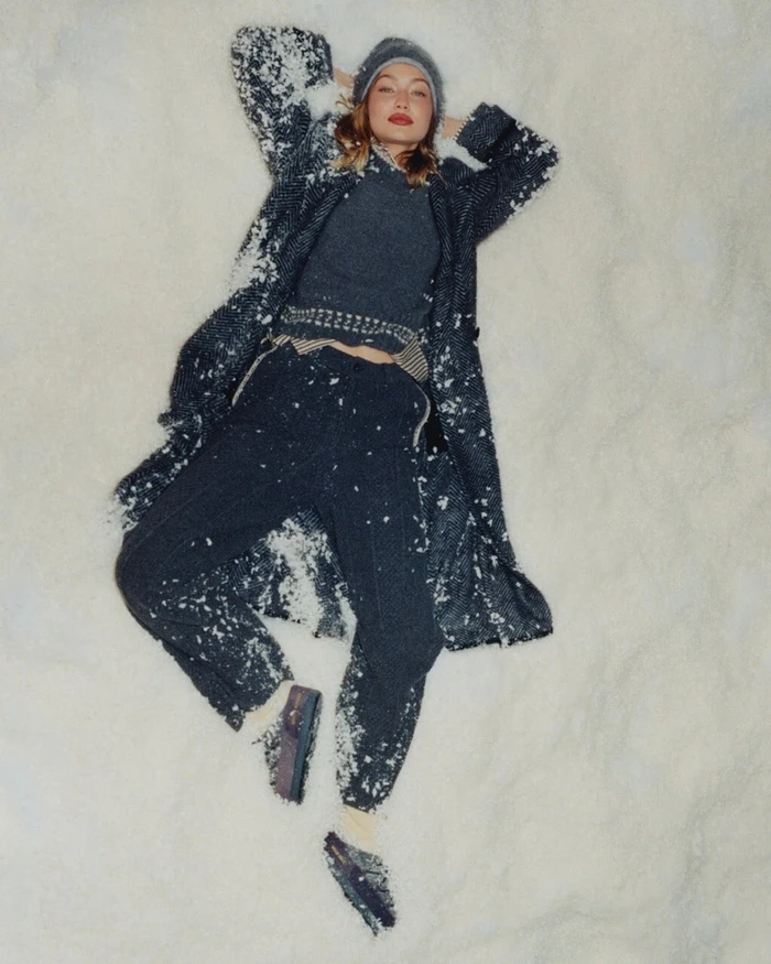 Gigi Hadid lying in snow wearing a dark grey knit coat, sweater, trousers, beanie and loafers.