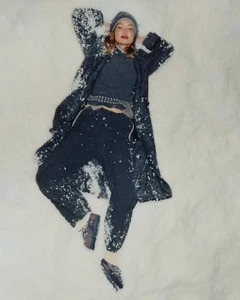 Gigi Hadid lying in snow wearing a dark grey knit coat, sweater, trousers, beanie and loafers.