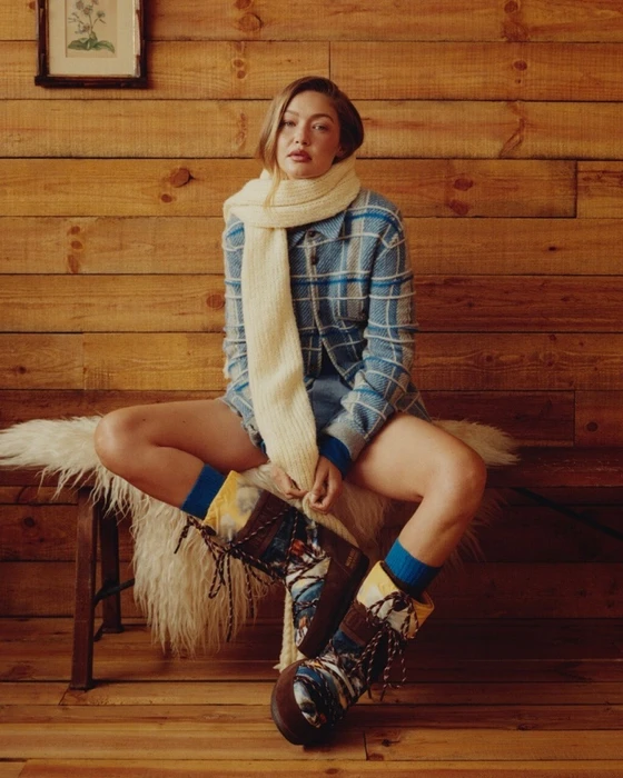 Gigi Hadid wearing a blue plaid shirt and cream scarf seated on a wooden bench against wood-paneled walls.