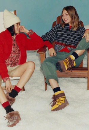 Two women seated in wooden lounge chairs on a white snow setting wearing winter apparel including knit sweaters, boots, and a beanie.