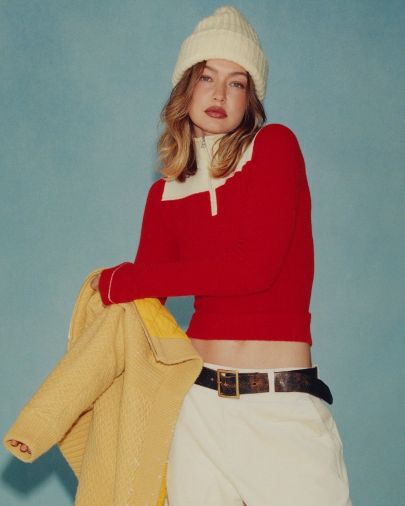 Model Gigi Hadid wearing a red knit sweater, white beanie, and holding a yellow jacket against a blue background.